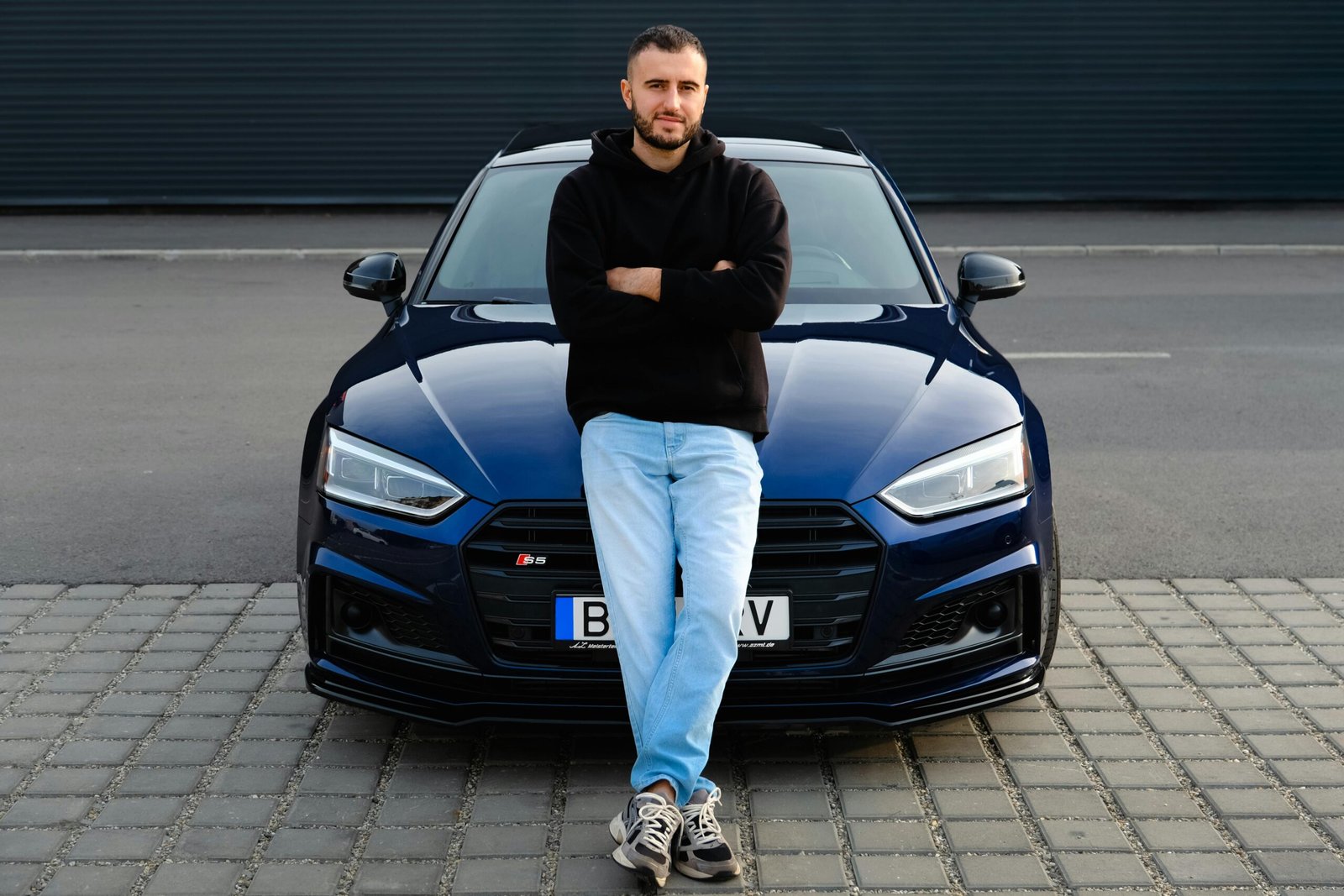 Man casually leaning on a blue car on a street, displaying urban fashion and style.